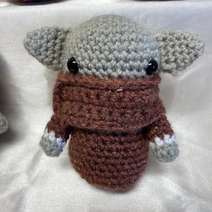 May include: A crocheted gray and brown character with large ears and a brown mask. The character has black eyes and a crocheted texture.