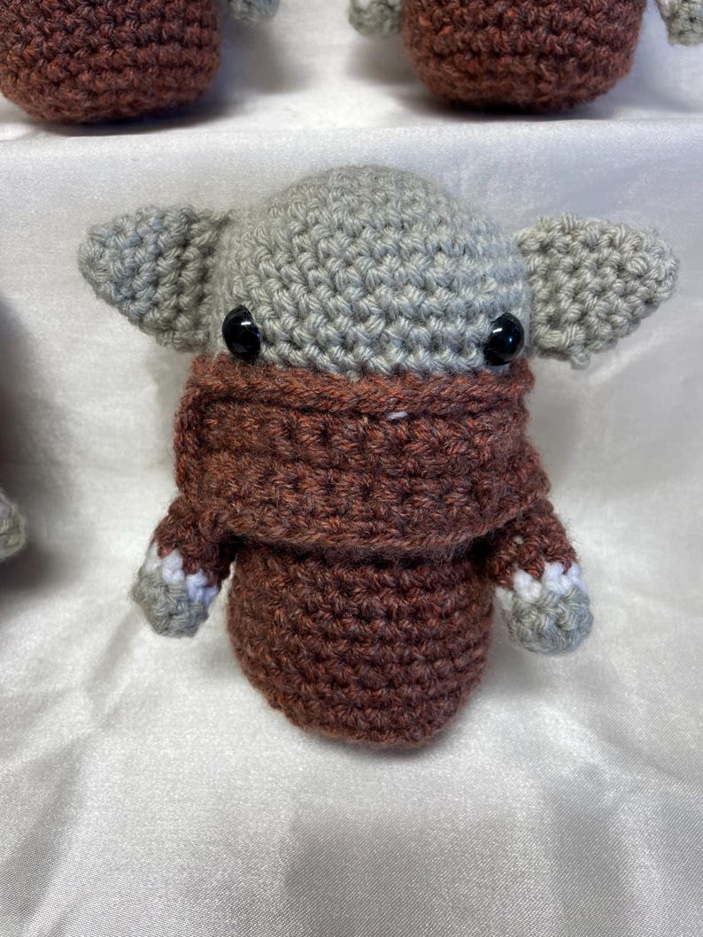 May include: A crocheted gray and brown character with large ears and a brown mask. The character has black eyes and a crocheted texture.