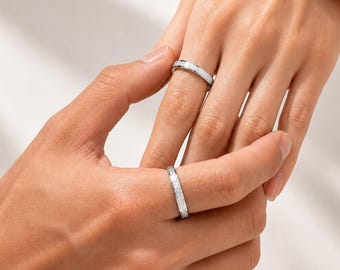 Könnte beinhalten: Zwei silberfarbene Ringe mit einer texturierten, matten Oberfläche. Jeder Ring hat ein schmales, gleichmäßiges Banddesign. Die Metalloberfläche weist eine subtile, körnige Struktur auf, die das Licht einfängt und einen Kontrast zu den glatten Kanten bildet.
