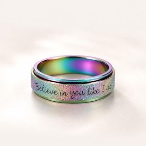 May include: A rainbow-colored spinner ring with a textured, iridescent finish. The outer band features the engraved black script text "Believe in you like I do" wrapped around the circumference of the metal ring, showcasing vibrant color shifts.