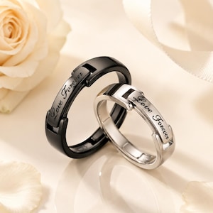 May include: Two rings, one black and one silver, each featuring a hinged design element on the band. The text "Love Forever" is engraved in a cursive script on the flat, rectangular section of each ring. The rings are positioned together.