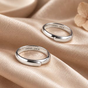 Op de afbeelding: Twee gepolijste zilverkleurige ringen met een glad, afgerond bandontwerp. Elke ring heeft een open stijl met overlappende uiteinden en is gegraveerd met de tekst EMMA JACK aan de binnenzijde van de band, wat een metalen afwerking vertoont.