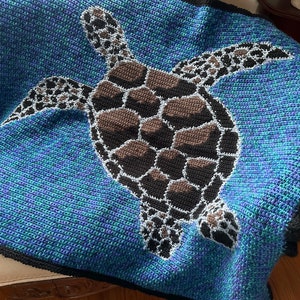 Puede incluir: Una manta de ganchillo con un diseño de tortuga marina. La tortuga es marrón y negra con detalles blancos y está nadando sobre un fondo azul y verde.