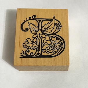 May include: A wooden stamp with a black, ornate letter "B" design. The letter is embellished with floral and leaf motifs, set against a light-colored wood block. The stamp is rectangular and appears unused.