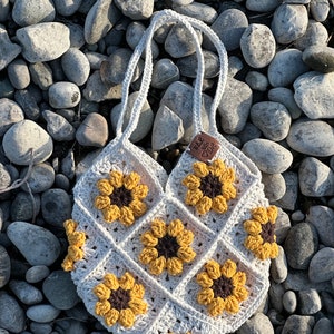 Könnte beinhalten: Eine weiße gehäkelte Tragetasche mit einem braunen Lederanhänger, auf dem "Made with Love" steht. Die Tasche ist mit gehäkelten Sonnenblumen in Gelb und Braun verziert.