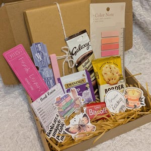 May include: A brown cardboard gift box filled with reading-themed items. Includes a pink bookmark, Galaxy instant chocolate, Twinings tea, a Biscoff cookie, stickers, and a notepad. The box is filled with shredded paper.