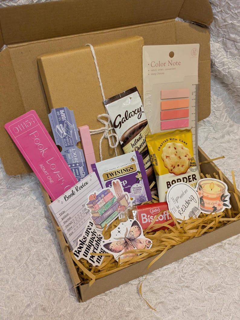 May include: A brown cardboard gift box filled with reading-themed items. Includes a pink bookmark, Galaxy instant chocolate, Twinings tea, a Biscoff cookie, stickers, and a notepad. The box is filled with shredded paper.
