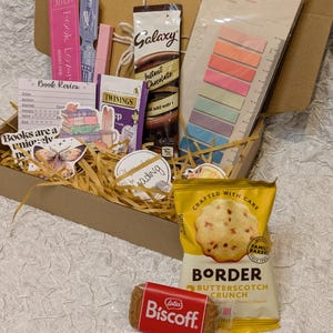 May include: A gift box filled with reading-themed items. Includes a Galaxy instant chocolate packet, Twinings tea, a pack of Border Butterscotch Crunch biscuits, a Lotus Biscoff biscuit, sticky notes, and bookmarks. The box is filled with straw.