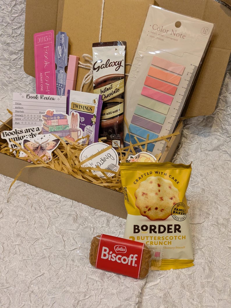 May include: A gift box filled with reading-themed items. Includes a Galaxy instant chocolate packet, Twinings tea, a pack of Border Butterscotch Crunch biscuits, a Lotus Biscoff biscuit, sticky notes, and bookmarks. The box is filled with straw.