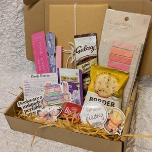 May include: A gift box filled with reading-themed items. Includes a book, snacks, tea, sticky notes, a bookmark, and stickers. The box is brown, and the contents are arranged on a bed of straw.