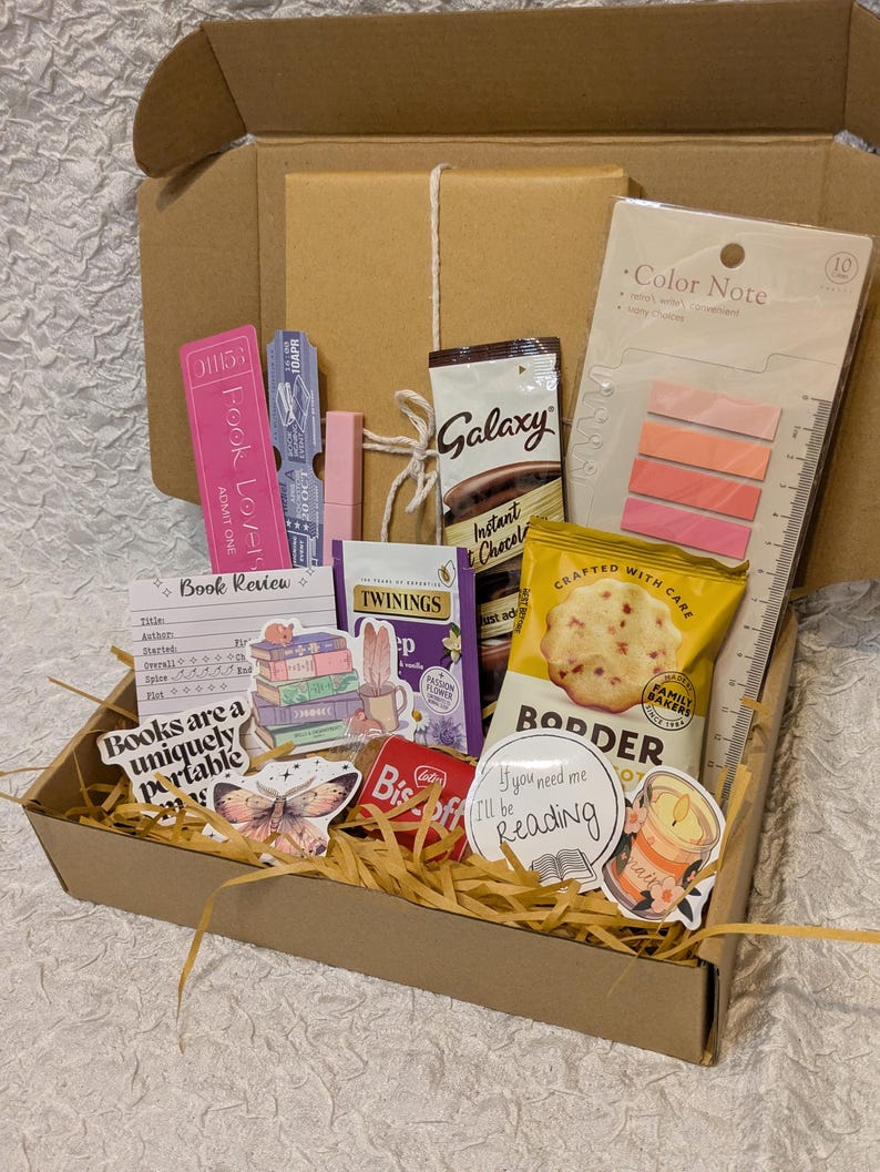 May include: A gift box filled with reading-themed items. Includes a book, snacks, tea, sticky notes, a bookmark, and stickers. The box is brown, and the contents are arranged on a bed of straw.