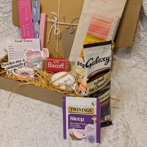 May include: A brown cardboard box filled with various items, including bookmarks, stickers, a Lotus Biscoff cookie, and a packet of Galaxy Instant Hot Chocolate. The box also contains a packet of Twinings Sleep tea and a Color Note sticky note pad.