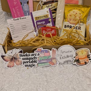 May include: A gift box filled with book-themed items. Includes a pink bookmark, a book review card, tea, chocolate, cookies, and stickers with book-related quotes and illustrations. The box is filled with decorative straw.