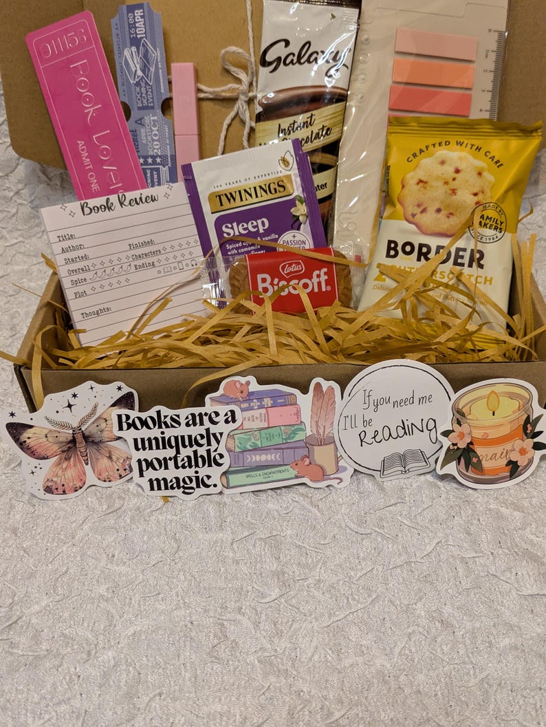 May include: A gift box filled with book-themed items. Includes a pink bookmark, a book review card, tea, chocolate, cookies, and stickers with book-related quotes and illustrations. The box is filled with decorative straw.