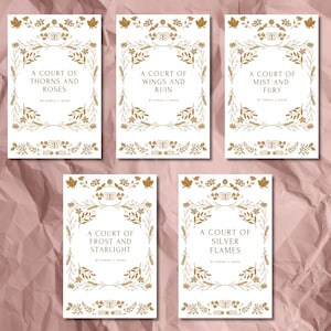 Puede incluir: Cinco libros con cubiertas blancas y diseños florales dorados. Cada libro tiene un título en una fuente decorativa, como "A Court of Thorns and Roses". El nombre del autor, Sarah J. Maas, está debajo. Los libros están sobre una superficie rosa.