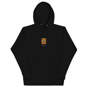 Puede incluir: Sudadera con capucha negra con una pequeña imagen bordada de un waffle con cara y un ojo. El waffle es naranja y marrón.