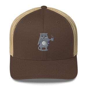 Puede incluir: Una gorra de camionero marrón y beige con un gato gris que sostiene una taza de café bordada en la parte delantera.