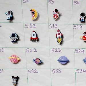 May include: A grid of 50 small, colourful, plastic charms with space-themed designs. The charms include astronauts, rockets, planets, stars, and other space-related imagery.
