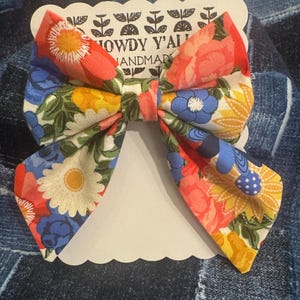May include: A large floral print bow with a red, yellow, blue, and white color scheme. The bow is tied with a white ribbon and has the text "Howdy Y'all Handmade" on a white tag.
