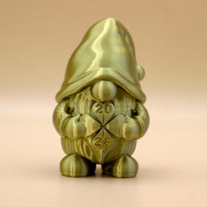 Könnte beinhalten: Goldfarbene Zwergenfigur mit spitzer Mütze und Kleeblatt-Design auf der Brust. Die Zahlen "20" und "25" sind auf dem Kleeblatt zu sehen. Die Figur hat eine strukturierte Oberfläche und ein skurriles Design.