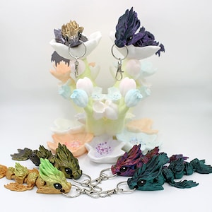 Könnte beinhalten: Ein Set mit sechs bunten Drachen-Schlüsselanhängern. Die Drachen sind aus Harz gefertigt und haben verschiedene Farben, darunter Gold, Grün, Lila und Blau. Die Schlüsselanhänger sind an silbernen Schlüsselringen befestigt.