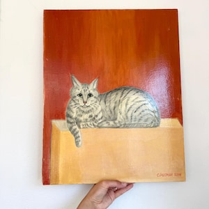 May include: An original painting of a gray tabby cat resting on a yellow surface against a red-orange background. The cat is painted with detailed stripes and is looking at the viewer. The painting is signed "C. Putman 5109".