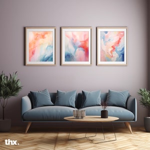 Può includere: Tre dipinti astratti ad acquerello in un soggiorno. I dipinti sono incorniciati in legno e presentano una miscela di tonalità rosa, blu e arancio. I dipinti sono appesi a una parete di colore lilla chiaro sopra un divano blu con tre cuscini. Un tavolino da caffè in legno con un vaso di vetro si trova di fronte al divano.