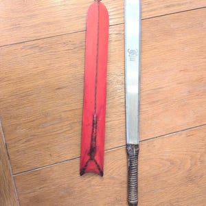 May include: A long, straight metal blade with a rounded tip and a dark, textured handle, accompanied by a separate, bright red, elongated sheath featuring a stitched seam and a small metal cap at the top end, displayed on a wooden surface.