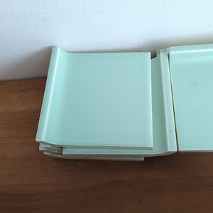 May include: A stack of light teal-colored square tiles with rounded edges. The tiles are stacked on a wooden surface, with a white wall in the background. The tiles appear to be new and unused.