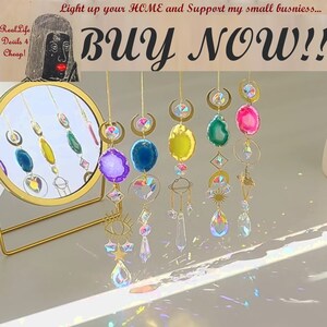 May include: Six iridescent crystal suncatchers hanging from gold chains. Each suncatcher features a different crystal shape and design, including a crescent moon, a star, an eye, and a heart. The suncatchers are hanging in front of a mirror with a gold frame.