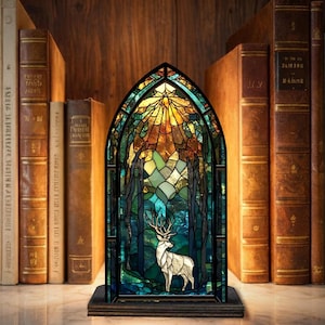 Könnte beinhalten: Eine Buntglaslampe mit einer Waldszene und einem weißen Hirsch. Die Lampe hat einen schwarzen Rahmen und einen Holzsockel. Im Hintergrund eine Sammlung antiker Bücher.