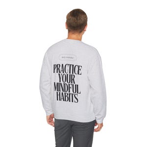 May include: Gray sweatshirt with long sleeves and a crew neck. The back of the sweatshirt has the text "daily reminder" in a small oval and the text "PRACTICE YOUR MINDFUL HABITS" in large black letters.
