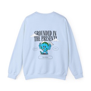 May include: Light blue sweatshirt with a cartoon illustration of a smiling earth with arms and legs. The text "GROUNDED IN THE PRESENT!" is printed above the illustration. The words "GROWING" and "WITH" are printed in smaller text on either side of the illustration. The text "THE WORLD" is printed below the illustration.