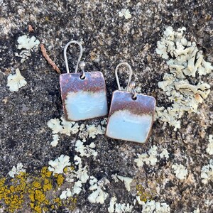 May include: A pair of square drop earrings featuring a two-tone design with a smooth, pale blue lower section and a textured, metallic brown upper section. The earrings are suspended from silver-toned metal ear wires with a simple hook closure.