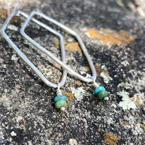 May include: A pair of silver-toned earrings featuring elongated hexagonal frames. Each earring is accented with two small, round, turquoise-colored beads and a tiny silver-toned bead at the base, attached to the bottom of the metal frame.