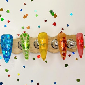 Könnte beinhalten: Sechs transparente Nagelspitzen mit verschiedenen farbigen, herzförmigen Glitzerdesigns. Die Spitzen sind auf einer weißen Oberfläche mit einer Holzbasis angeordnet.