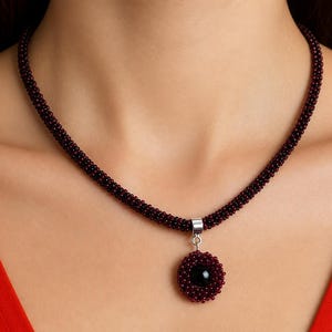 Könnte beinhalten: Eine dunkelweinrote Perlenkette mit einem kreisförmigen Anhänger, der eine schwarze Perle in der Mitte hat. Die Kette besteht aus zahlreichen winzigen, gleichmäßigen dunkelroten Perlen, die zu einer zylindrischen Schnur gewebt sind.