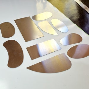 May include: A collection of various metal palette knives and scrapers in different shapes and sizes. The tools are arranged on a white surface, with some showing a brushed metal finish and others a copper-toned appearance. A metal ruler is visible in the background.