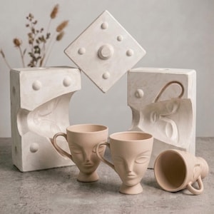 Può includere: Una natura morta con tre tazze in ceramica beige con volti scolpiti, una quarta tazza sdraiata su un lato e uno stampo. Le tazze hanno manici e sono su una superficie grigia. Lo sfondo include un vaso con fiori secchi.