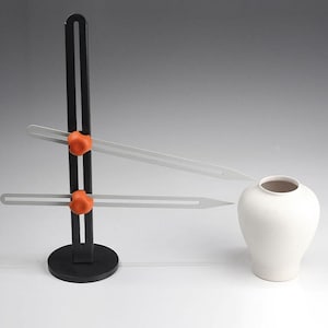 Op de afbeelding: Een zwart pottenbakkerswielgereedschap met twee verstelbare armen en oranje knoppen. Het gereedschap wordt gebruikt om symmetrische vormen op aardewerk te creëren.