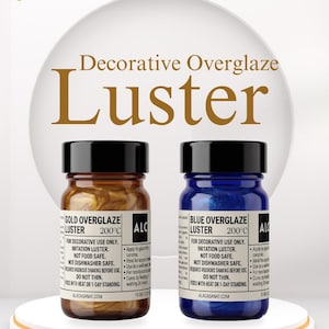 May include: Two small glass bottles of decorative overglaze luster, one gold and one blue, with black caps. The bottles are labeled with instructions and the brand name. The background is white with gold confetti.