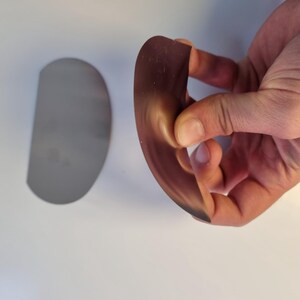 May include: A curved, semi-circular, brown object is held in a hand, with a similar, flat, silver-colored object to the left. The brown object appears flexible, with a gradient from light to dark. The objects are on a white surface.