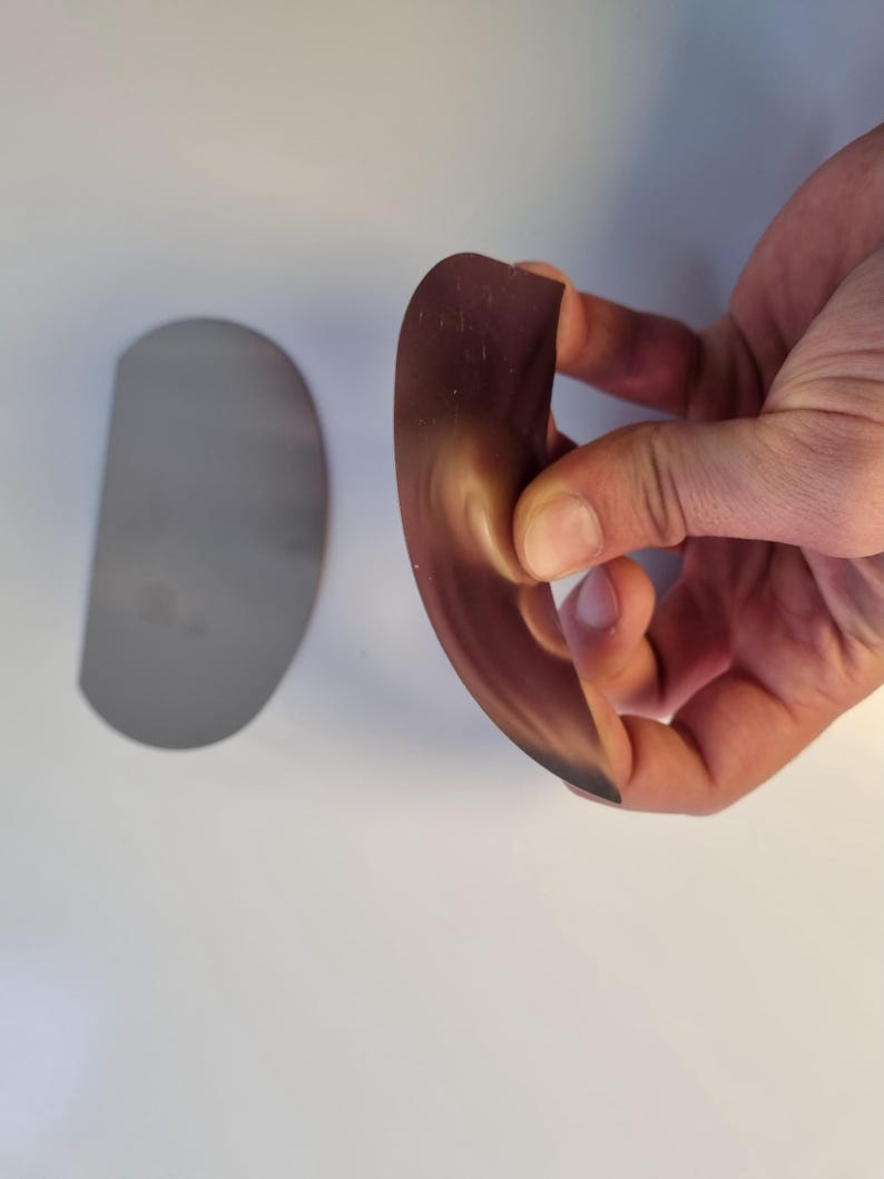 May include: A curved, semi-circular, brown object is held in a hand, with a similar, flat, silver-colored object to the left. The brown object appears flexible, with a gradient from light to dark. The objects are on a white surface.