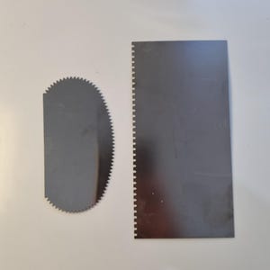 May include: Two stainless steel cake decorating tools on a white surface. One is a semi-circular serrated edge scraper, and the other is a rectangular scraper with a serrated edge on one side. These tools are used for smoothing frosting.