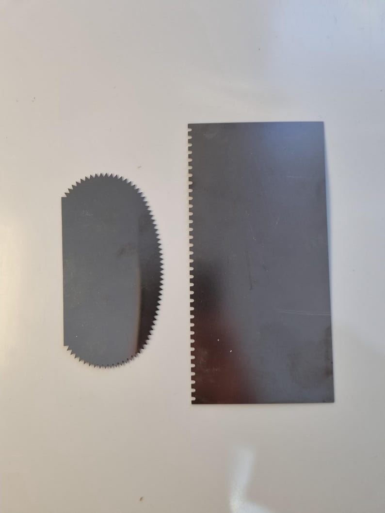 May include: Two stainless steel cake decorating tools on a white surface. One is a semi-circular serrated edge scraper, and the other is a rectangular scraper with a serrated edge on one side. These tools are used for smoothing frosting.