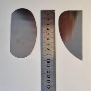 May include: Two silver-colored, semi-circular metal objects next to a metal ruler. The ruler is marked with millimeter increments and measures up to 18 cm. The objects are on a white surface.