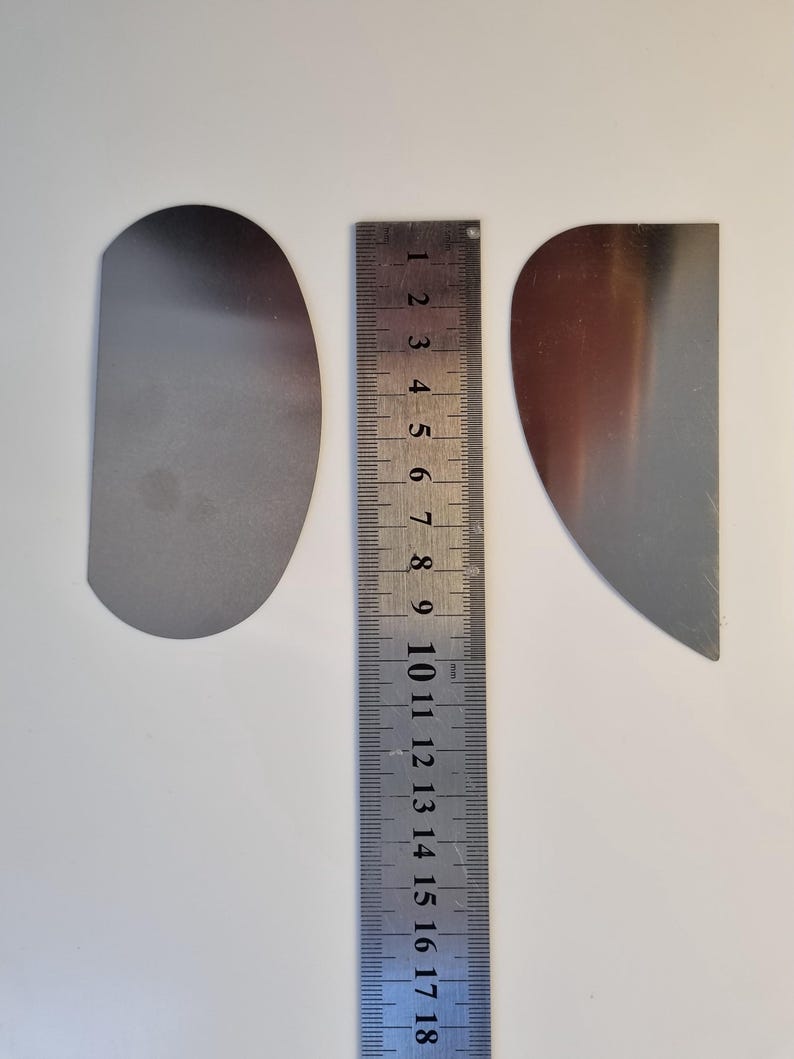 May include: Two silver-colored, semi-circular metal objects next to a metal ruler. The ruler is marked with millimeter increments and measures up to 18 cm. The objects are on a white surface.