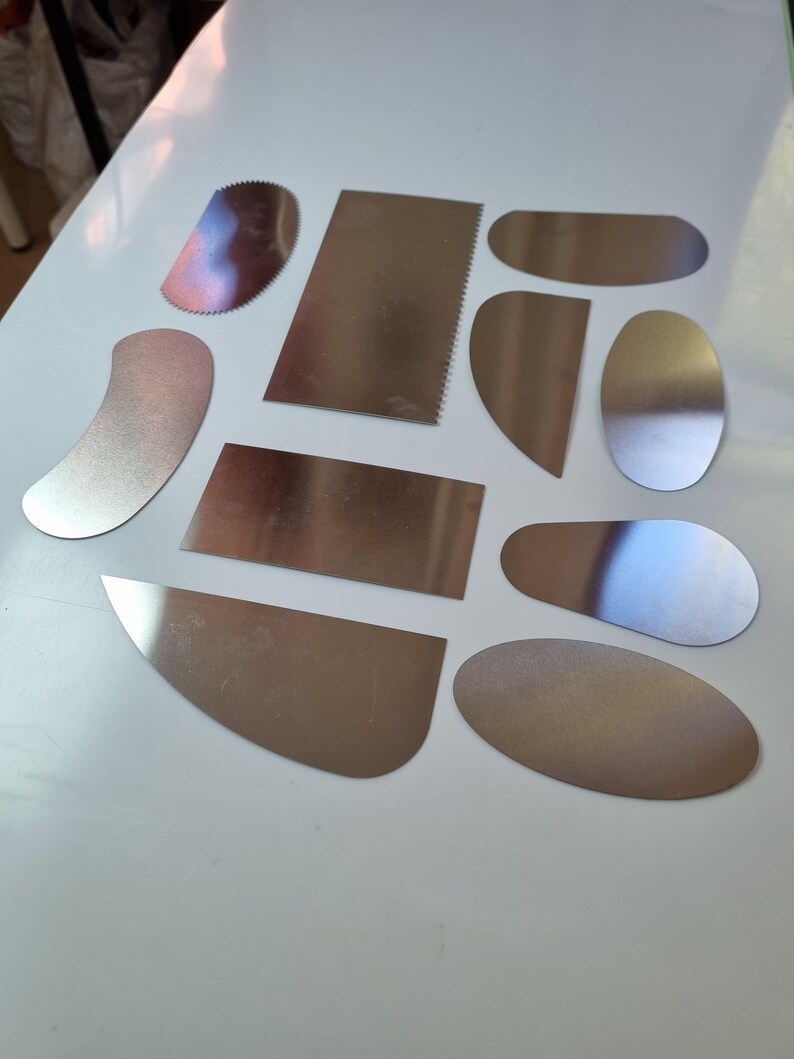 May include: A collection of various stainless steel pottery tools, including curved, straight, and serrated edge shapes. The tools are arranged on a white surface, reflecting light and showcasing their metallic finish. These tools are designed for shaping and smoothing clay.