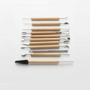 Può includere: Un set di strumenti per la ceramica con manici in legno e punte in metallo. Gli strumenti includono varie forme come anelli, aghi e spazzole. Gli strumenti sono disposti in file parallele su uno sfondo bianco.