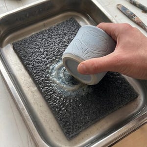Op de afbeelding: Een lichtblauwe keramische beker met een bloemenontwerp wordt schoongemaakt. De beker wordt boven een donkergrijze, getextureerde spons in een roestvrijstalen bak met water gehouden. Pottenbakken en keramiek.